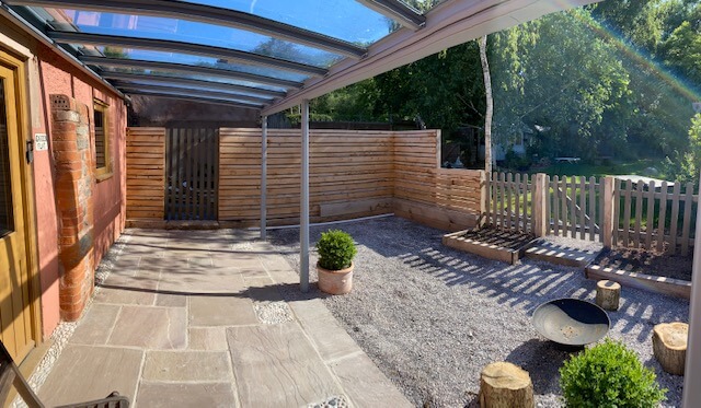 Patio area featuring a glass canopy, gravel surface, wooden fence, and potted plants.