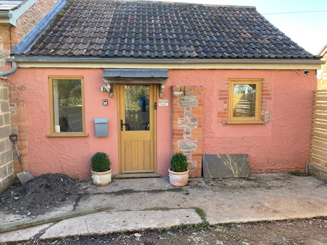 Cottage featuring a pink exterior, wooden door, and two windows with potted plants on either side.