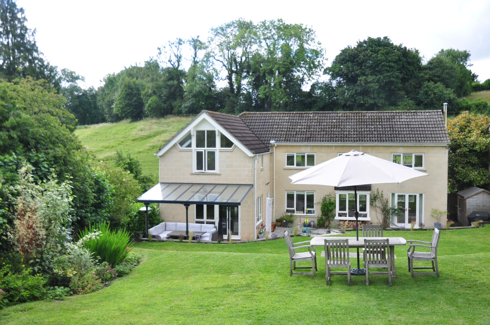 Two-storey house with a veranda and garden furniture in a green outdoor setting.