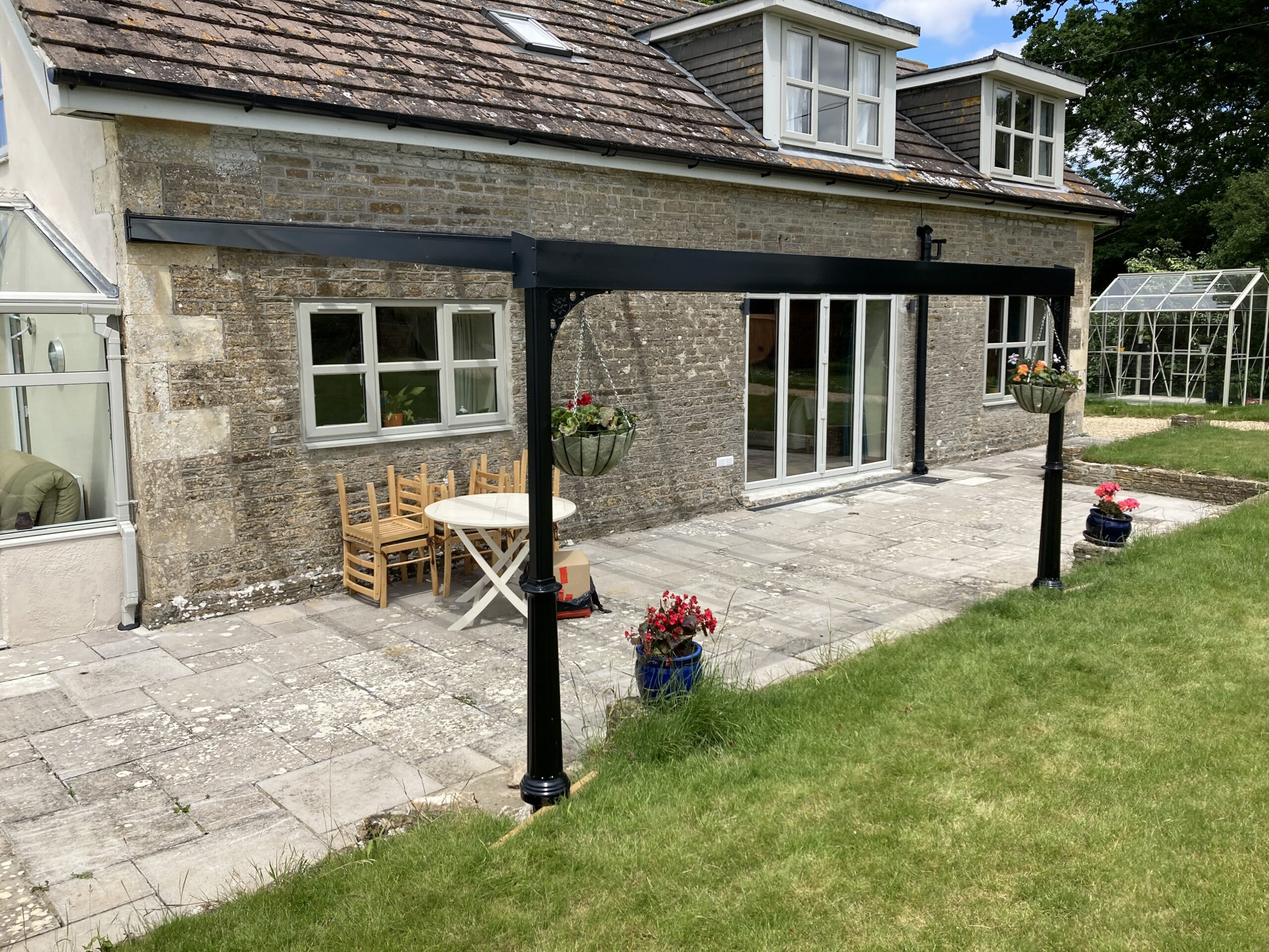 Jet black glass roof Victorian canopy structure with hanging planters in a garden setting.