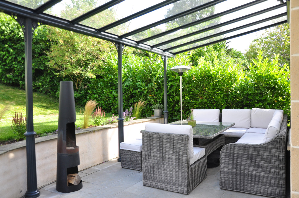Veranda canopy with a glass roof in anthracite grey, providing shelter over a seating area.