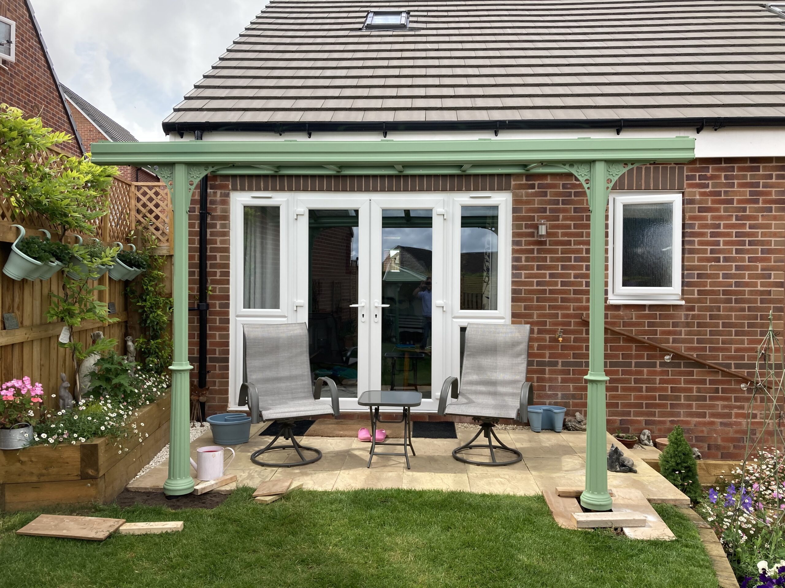 Pale green veranda canopy with a glass roof supported by decorative columns in a garden setting.